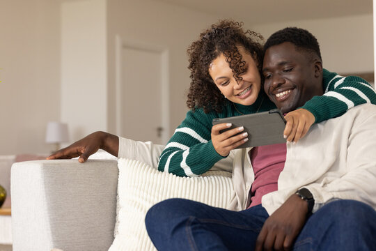 African American couple sitting closely on light colored couch, leaning over tablet in living room