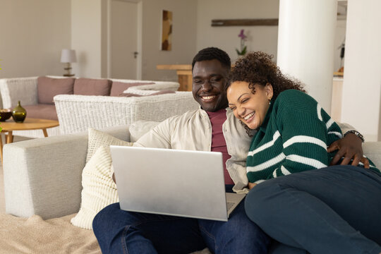 African American couple sitting on sofa, smiling and watching laptop in cozy living room