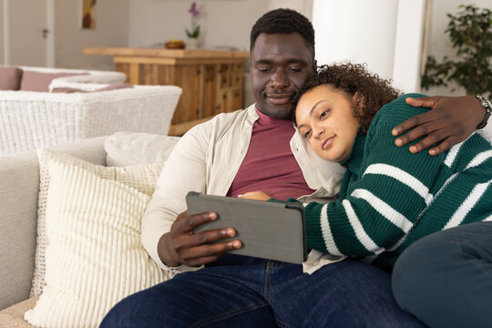 African American couple sitting on sofa at home, holding tablet, wearing casual clothes and leaning