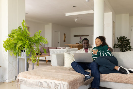 African couple sitting on sofa sharing silver laptop on man's lap, woman holding ceramic mug