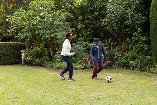 Father and teenage son kicking soccer ball on yard wearing hoodies and sneakers near planter