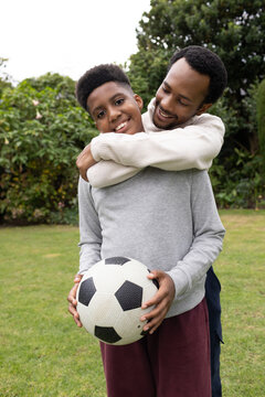 African American father and adolescent son standing on lawn, hugging while son holding soccer ball