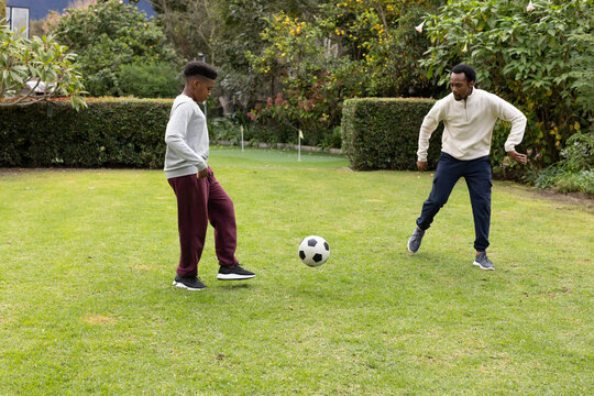 African American parent and teen males playing with soccer ball on backyard lawn near putting green