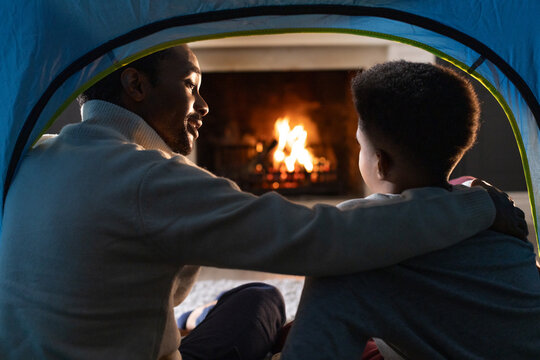 African American father and son sitting inside pop-up tent on carpeted floor, facing lit fireplace