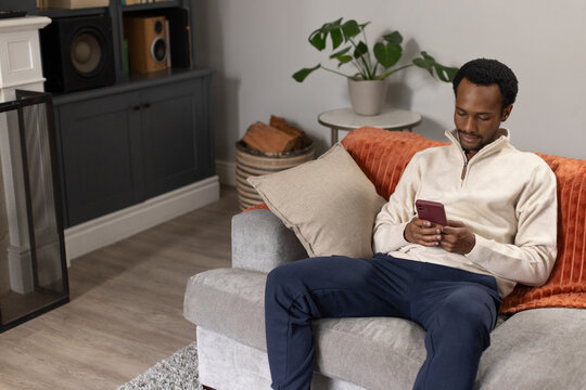 Adult male sitting on gray sofa in living room holding maroon smartphone, wearing beige pullover