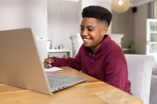 Teenage male student in maroon pullover sitting in living area writing with pencil using laptop