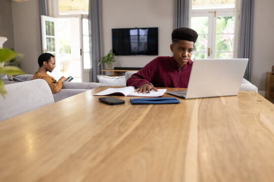 African American father and teenage son sitting at wooden table, son using laptop and notebook