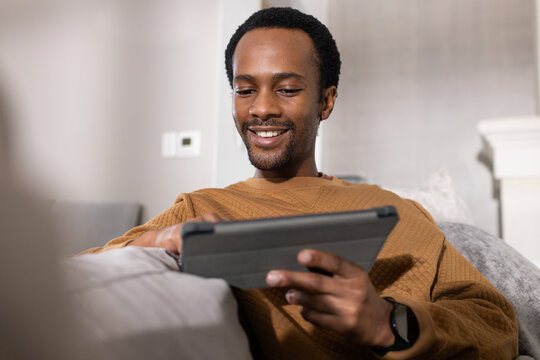 Man sitting on sofa at home using tablet folio case, wearing brown sweater and smartwatch, smiling