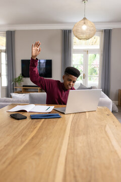 Teen African-American in burgundy sweater studying at home table using silver laptop, taking notes