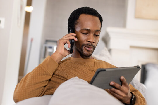 Adult African American man in brown sweater sitting on sofa at home using smartphone holding tablet