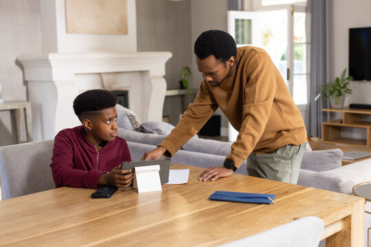 African American father and teen son studying at dining table in living room using tablet stand