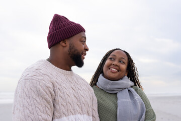 Obraz premium Couple standing facing sandy beach, wearing maroon beanie, knit sweaters, gray scarf, braids, sea