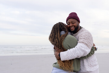 Obraz premium African American couple embracing, smiling on beach by ocean waves, wearing beanie and knit sweater