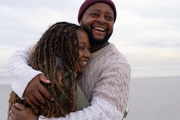 Obraz premium African American couple hugging, smiling on beach, maroon beanie, cable-knit, green sweater, braids