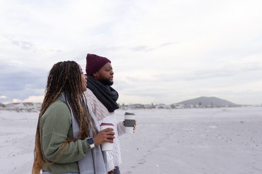 African couple standing on beach holding takeaway cups, wearing beanie and smartwatch, copy space