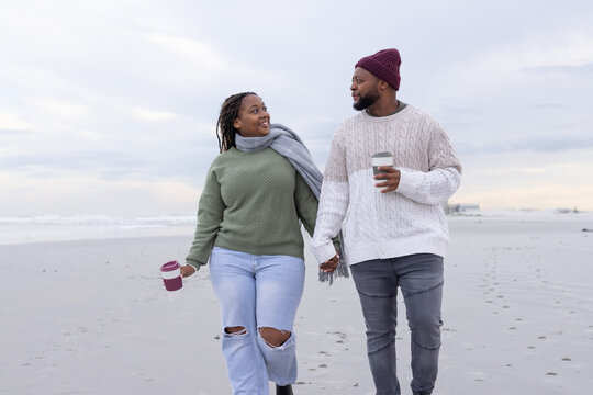 African American couple walking on beach in sweaters holding hands with coffee, leaving footprints