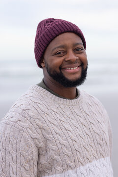 African American man in 30s smiling and standing on beach wearing maroon beanie, beige sweater