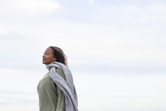 African woman standing under pale cloudy sky wearing olive knit sweater and grey shawl, copy space