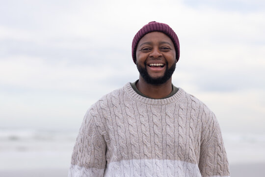 African American man standing on beach, smiling, wearing maroon beanie and cream cable-knit sweater