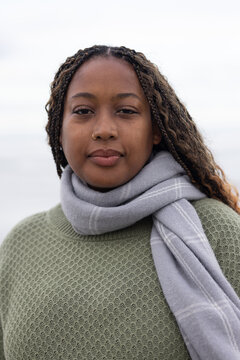 Woman standing facing camera at waterfront wearing olive sweater gray scarf nose ring braids