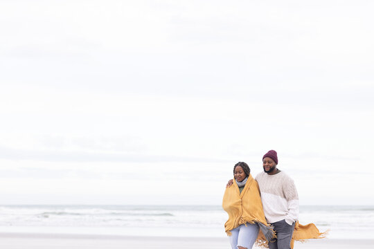 African couple walking arm in arm on beach, in mustard shawl and maroon beanie, copy space