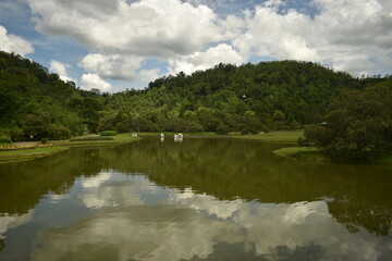 Obraz premium Scenic landscape of a calm lake reflecting fluffy white clouds and lush green rolling hills under a blue sky.