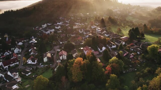 Flying over a scenic village and rural landscape with hills, meadows, woods, sunbeams and illuminated fog at sunrise.