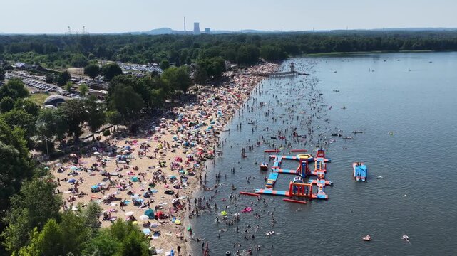Aerial drone view of a crowded summer lake beach with people swimming and sunbathing. Colorful umbrellas, water obstacle course on lake, on a sunny day. Lake Pogoria in Dabrowa Gornicza, Poland