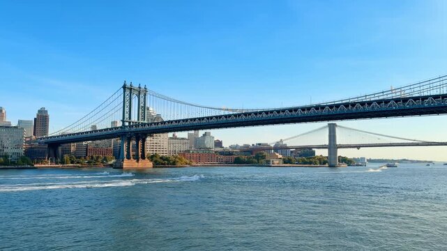 Manhattan Bridge spanning East River with Brooklyn Bridge beyond. Wide waterfront view of green suspension towers, wake trails and clear blue sky.