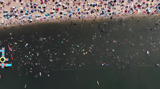 Aerial drone view of a crowded summer lake beach with people swimming and sunbathing. Colorful umbrellas, water obstacle course on lake, on a sunny day. Lake Pogoria in Dabrowa Gornicza, Poland