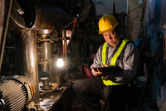Senior engineer using digital tablet to inspect machinery in low light environment with light bulb in factory.