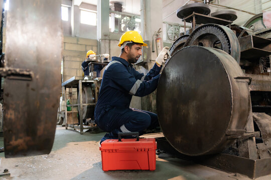 Technician wearing hardhat kneeling to inspect machine gear with red toolbox in industrial factory.