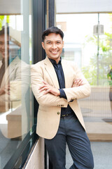 Fototapeta premium Smiling businessman in beige blazer standing with arms crossed by office glass wall, confidence, success, and a modern professional work setting.