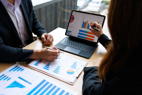 Two business professionals discussing sales charts and graphs on a laptop and paper reports during a team meeting at a desk in a modern office.