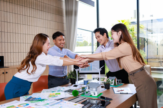 Four business colleagues stacking hands in a modern office, smiling over project plans and a small house model, showing teamwork, agreement and collaboration