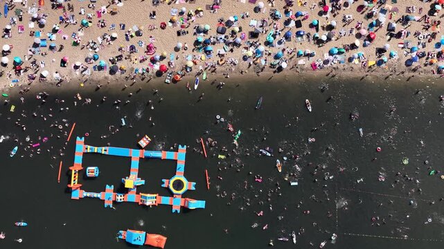Aerial drone view of a crowded summer lake beach with people swimming and sunbathing. Colorful umbrellas, water obstacle course on lake, on a sunny day. Lake Pogoria in Dabrowa Gornicza, Poland