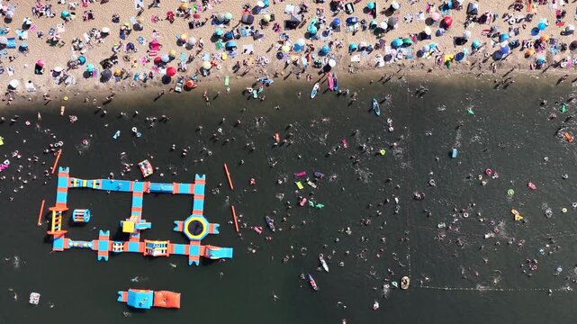 Aerial drone view of a crowded summer lake beach with people swimming and sunbathing. Colorful umbrellas, water obstacle course on lake, on a sunny day. Lake Pogoria in Dabrowa Gornicza, Poland