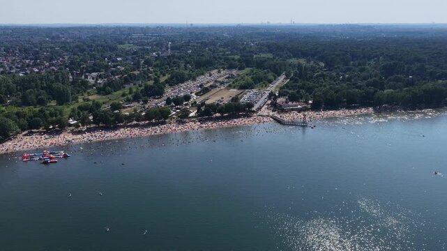 Aerial drone view of a crowded summer lake beach with people swimming and sunbathing. Colorful umbrellas, water obstacle course on lake, on a sunny day. Lake Pogoria in Dabrowa Gornicza, Poland
