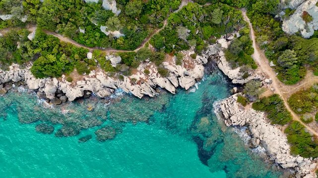 Aerial view of a beautiful turquoise sea next to a rocky coastline with lush green vegetation. The clear water reveals coral reefs, small waves gently wash the shore.  Sardinia holiday trip.