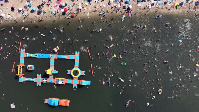 Aerial drone view of a crowded summer lake beach with people swimming and sunbathing. Colorful umbrellas, water obstacle course on lake, on a sunny day. Lake Pogoria in Dabrowa Gornicza, Poland