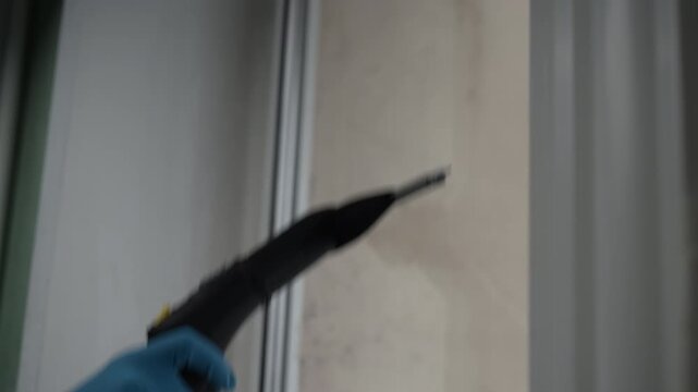 Close-up hand of professional janitor wearing protective gloves using powerful steam generator to remove stubborn dirt, mildew, mold from window frame, ensuring deep and hygienic clean for home.