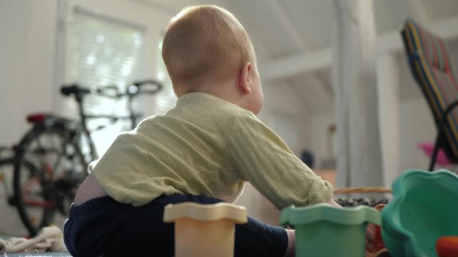 Baby in a light green sweater sits on a textured carpet toward the camera among scattered plastic toys and stacking cups