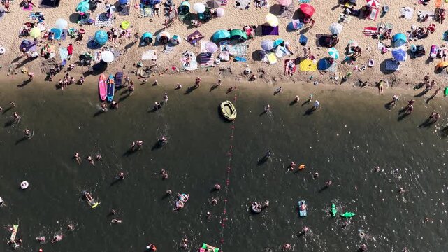 Aerial drone view of a crowded summer lake beach with people swimming and sunbathing. Colorful umbrellas, water obstacle course on lake, on a sunny day. Lake Pogoria in Dabrowa Gornicza, Poland