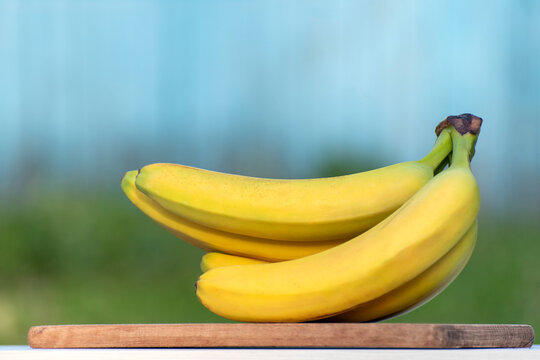 Bright yellow bananas are resting on a rustic wooden cutting board, presenting a fresh and wholesome fruit snack with a soft blue and green background