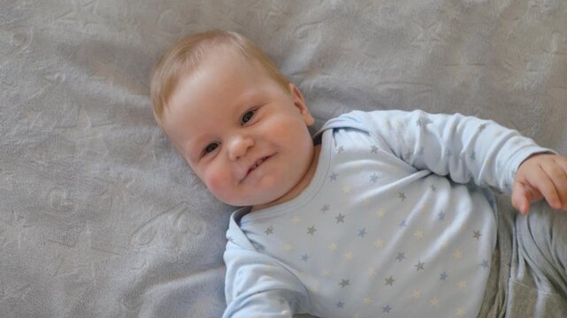 A happy baby waves his arms on a gray fleece blanket. The infant radiates positive emotions and smiles, top view