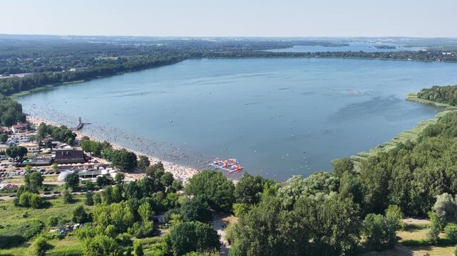 Aerial drone view of a crowded summer lake beach with people swimming and sunbathing. Colorful umbrellas, water obstacle course on lake, on a sunny day. Lake Pogoria in Dabrowa Gornicza, Poland