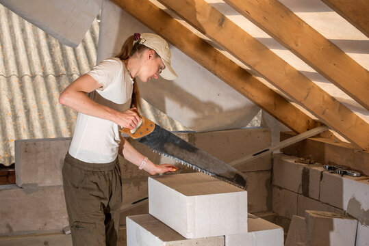 Female worker using a handsaw to cut a lightweight aerated concrete block during home renovation work on an attic construction site, focusing on efficiency and progress