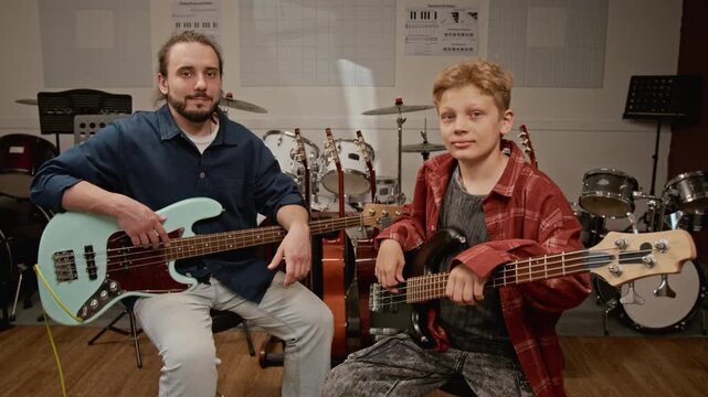 Portrait of young man and teenage boy holding bass guitars in music studio. Footag for promoting music education, private tutoring, and creative hobbies
