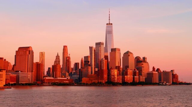 Lower Manhattan skyline glowing at sunset from Hudson River. Warm copper light reflects off glass towers with One World Trade Center rising above the horizon.