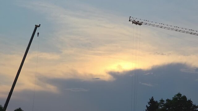 Construction cranes work in the evening sky as clouds gather and the sun sets behind them